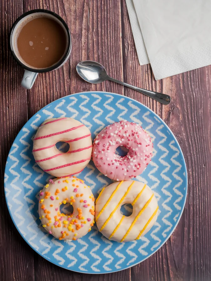 Cuatro donuts con coberturas de colores en plato azul junto a una taza sobre madera