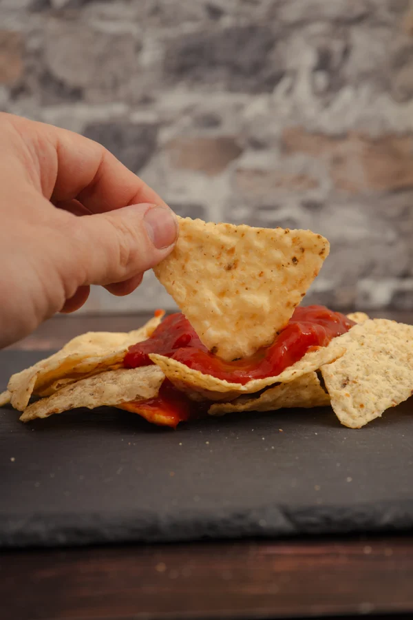 Una mano moja un nacho en salsa sobre una bandeja oscura