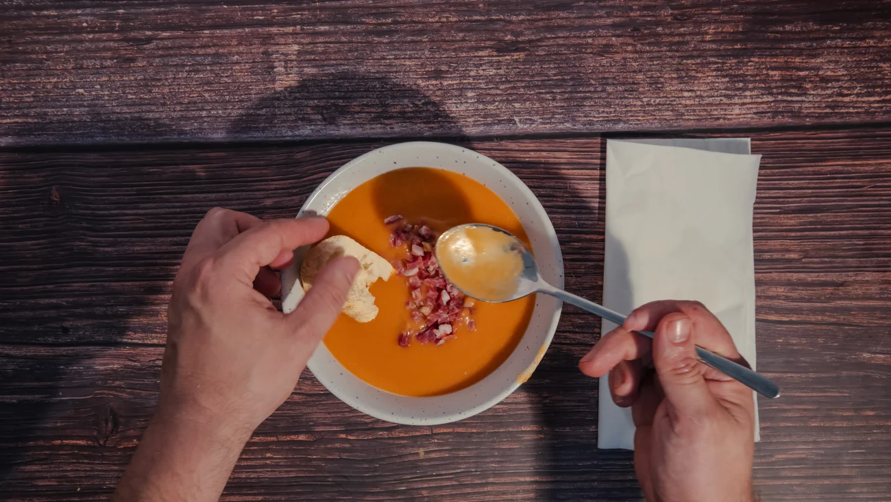 Manos compartiendo salmorejo con pan y cuchara sobre una mesa de madera